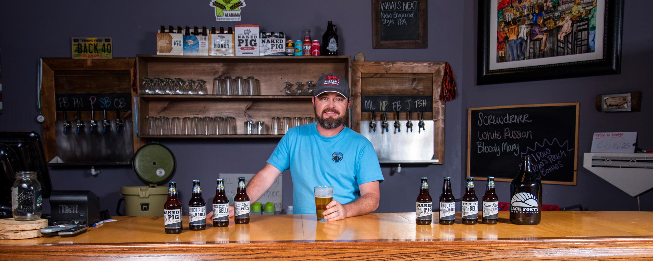 Photo of Jason Wilson at Back Forty brewery standing behind the bar with a glass up beer in his hand. 