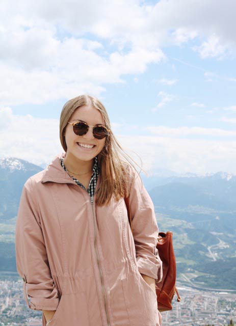 Jackie Ramirez smiling with sunglasses and a pink jacket on standing on top of a mountain.