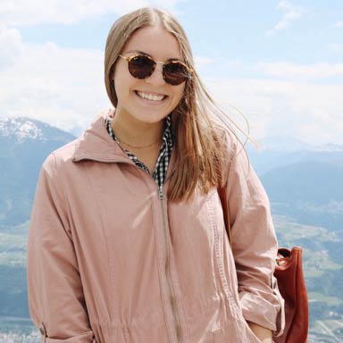 Jackie Ramirez smiling with sunglasses and a pink jacket on standing on top of a mountain.