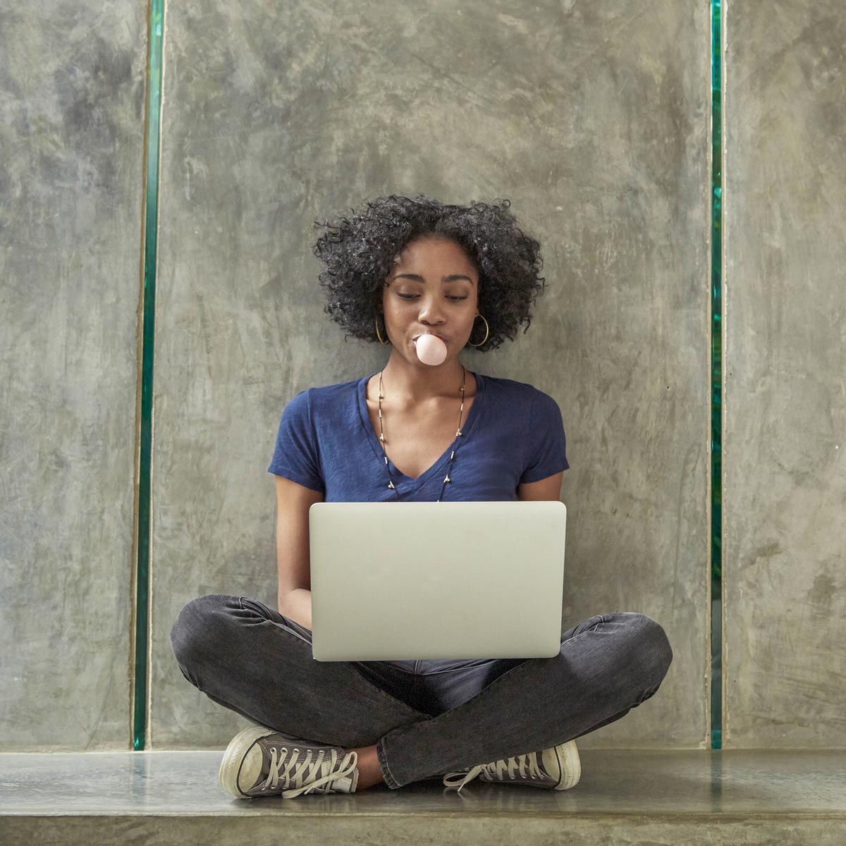 Female student sitting against a wall on a laptop.