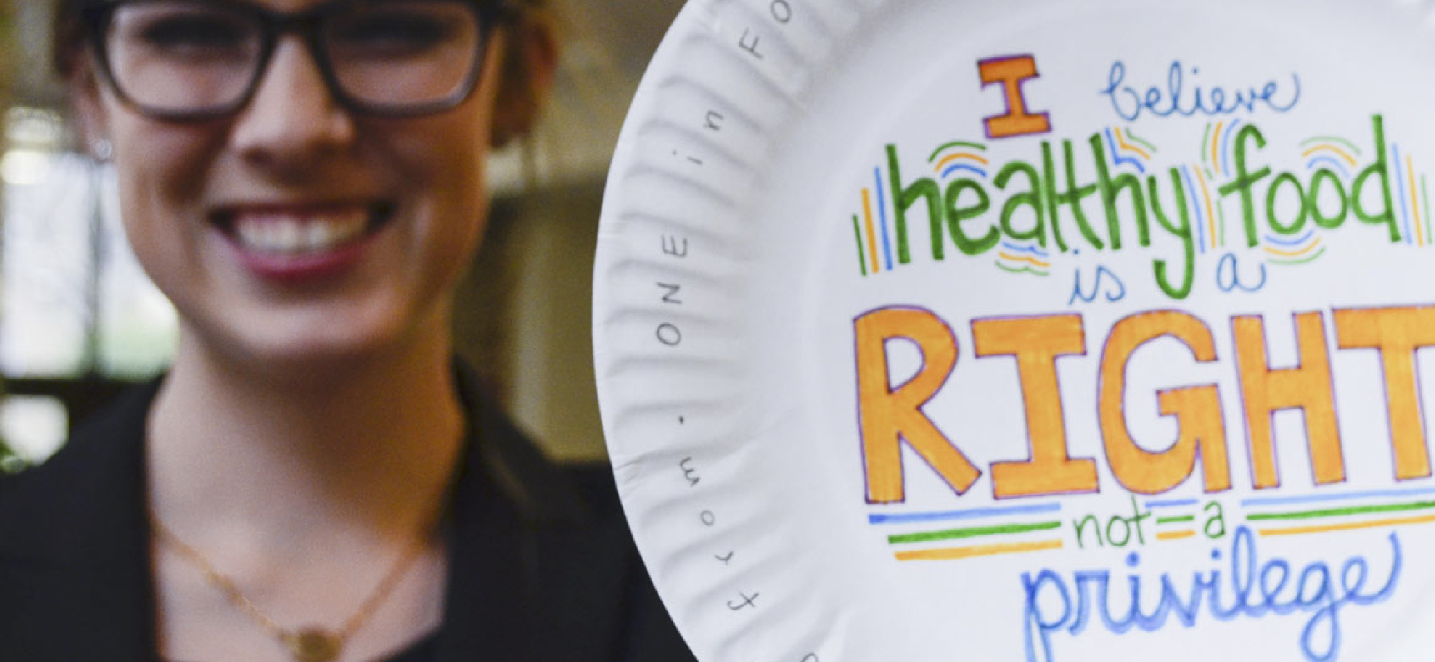 Photo of smiling female college student holding a paper plate with a handwritten message written in orange, blue and green marker that says 'I believe healthy food is a right not a privilege'.