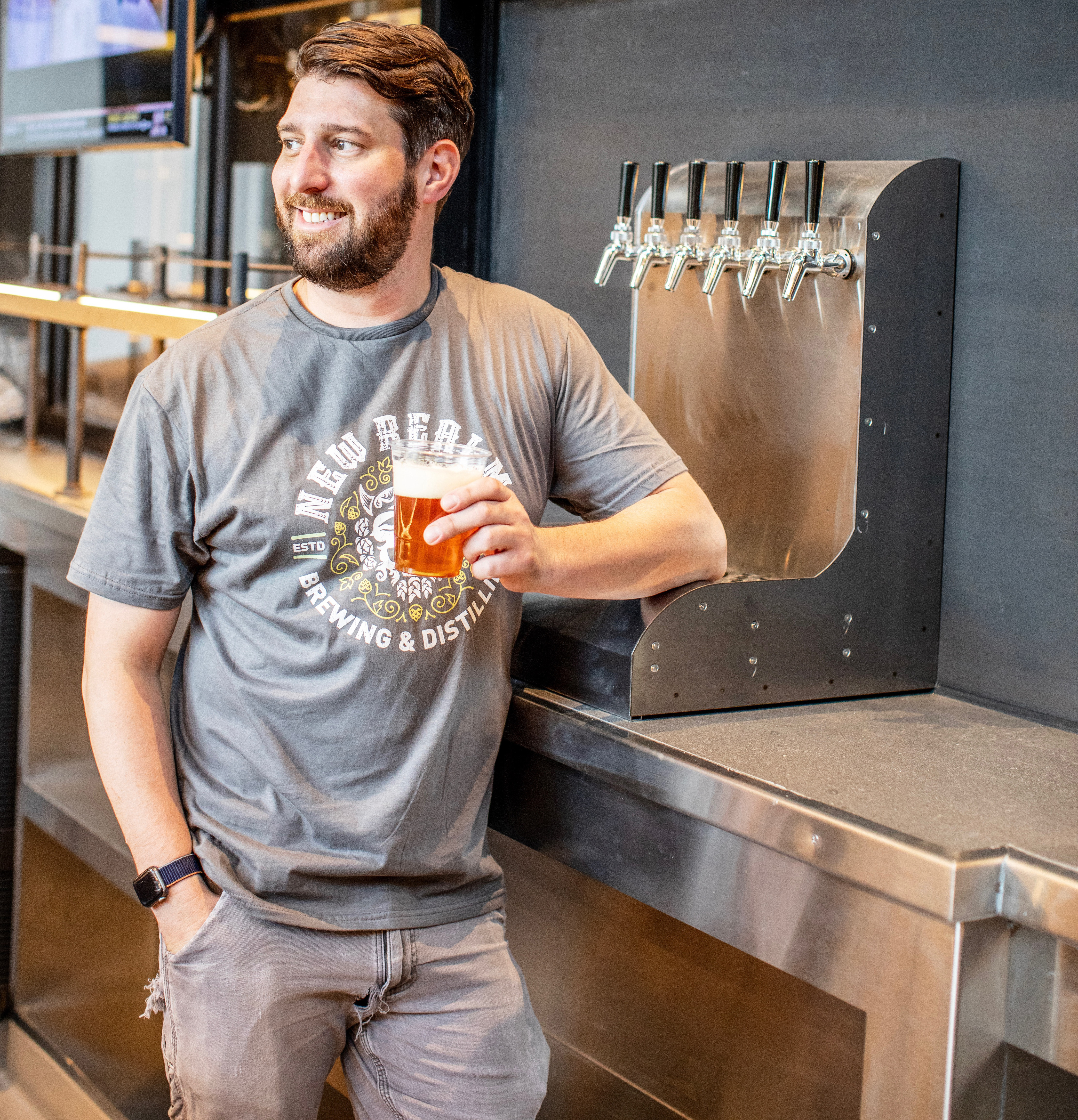 Drew Kostic leaning agaist the bar with a beer glass and breing tanks behind him.